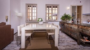 Uma área de jantar com mesa branca, cadeiras e um vaso central. Há duas janelas com molduras brancas e vista para edifícios. Um sofá marrom está encostado na parede, e um armário de madeira com espelho fica à direita.