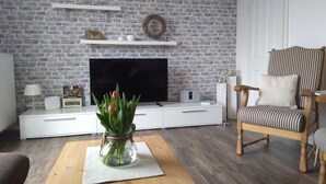 Una sala de estar con pared de ladrillo, un televisor de pantalla plana, un mueble blanco para entretenimiento, una guirnalda de mimbre, un sillón a rayas y una mesa de centro con un tarro de vidrio lleno de flores.