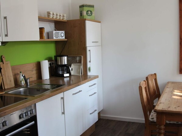 A compact kitchen with white cabinets, a coffee maker, a radio, and a wooden dining table.