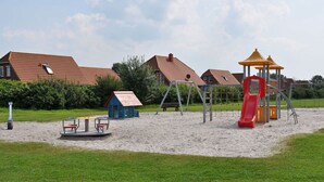 A playground with swings, a slide, and a climbing structure, surrounded by sand and grass, with houses in the background. - Beach Crab Modern Retreat (Dornum)
