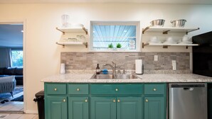 A kitchen with teal cabinets, a stainless steel sink, and a dishwasher. There are shelves above the sink holding dishes and a window with a view of greenery. - Modern in the Magic City (Irondale)