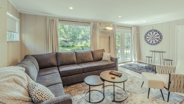 A modern living room with a sectional sofa, a coffee table, and a small side table. There is a large window with curtains, a dartboard on the wall, and a view of greenery outside. - Modern in the Magic City (Irondale)