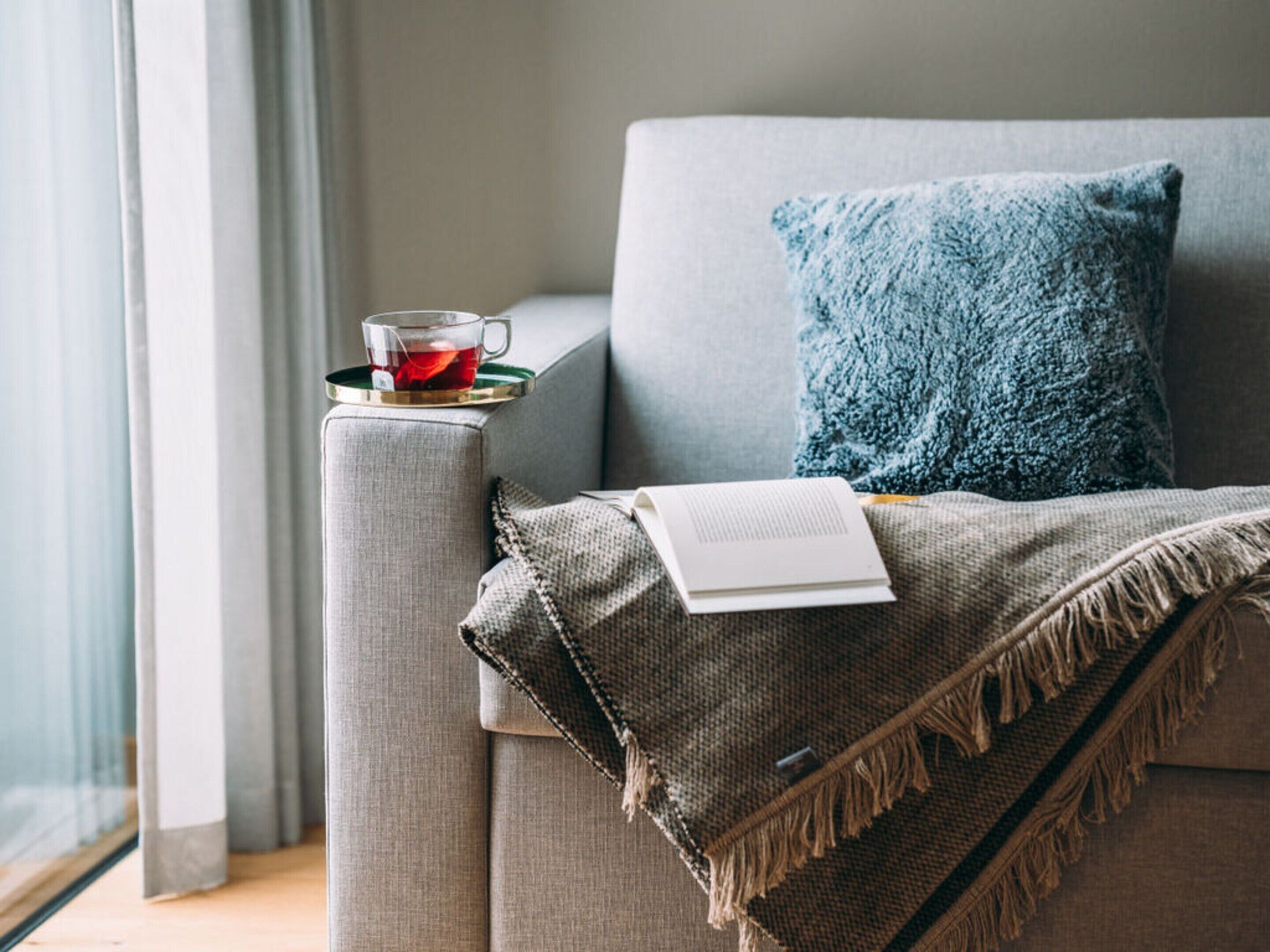 Un salon chaleureux avec un canapé gris, un coussin bleu, un verre d’une boisson rouge, un livre et une couverture en tissu tissé.