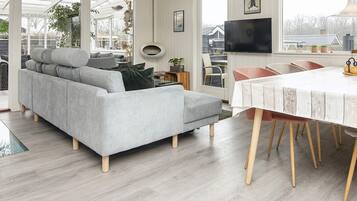 A modern living room with a grey sofa, a dining table with wooden chairs, and a large window with a view of the outdoors.