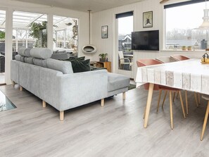 Un salon moderne avec un canapé gris, une table à manger aux pieds en bois et une grande fenêtre donnant sur l’extérieur.
