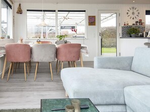 A modern living room with a grey sofa, a dining area with pink chairs, and a kitchen with a clock on the wall. - 5 Person Holiday Home in Juelsminde (Horsens)