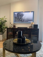 Una sala de estar con una mesa de centro redonda de mármol negro, un televisor de pantalla plana grande montado en la pared, una planta en maceta y una estantería con libros y objetos decorativos.