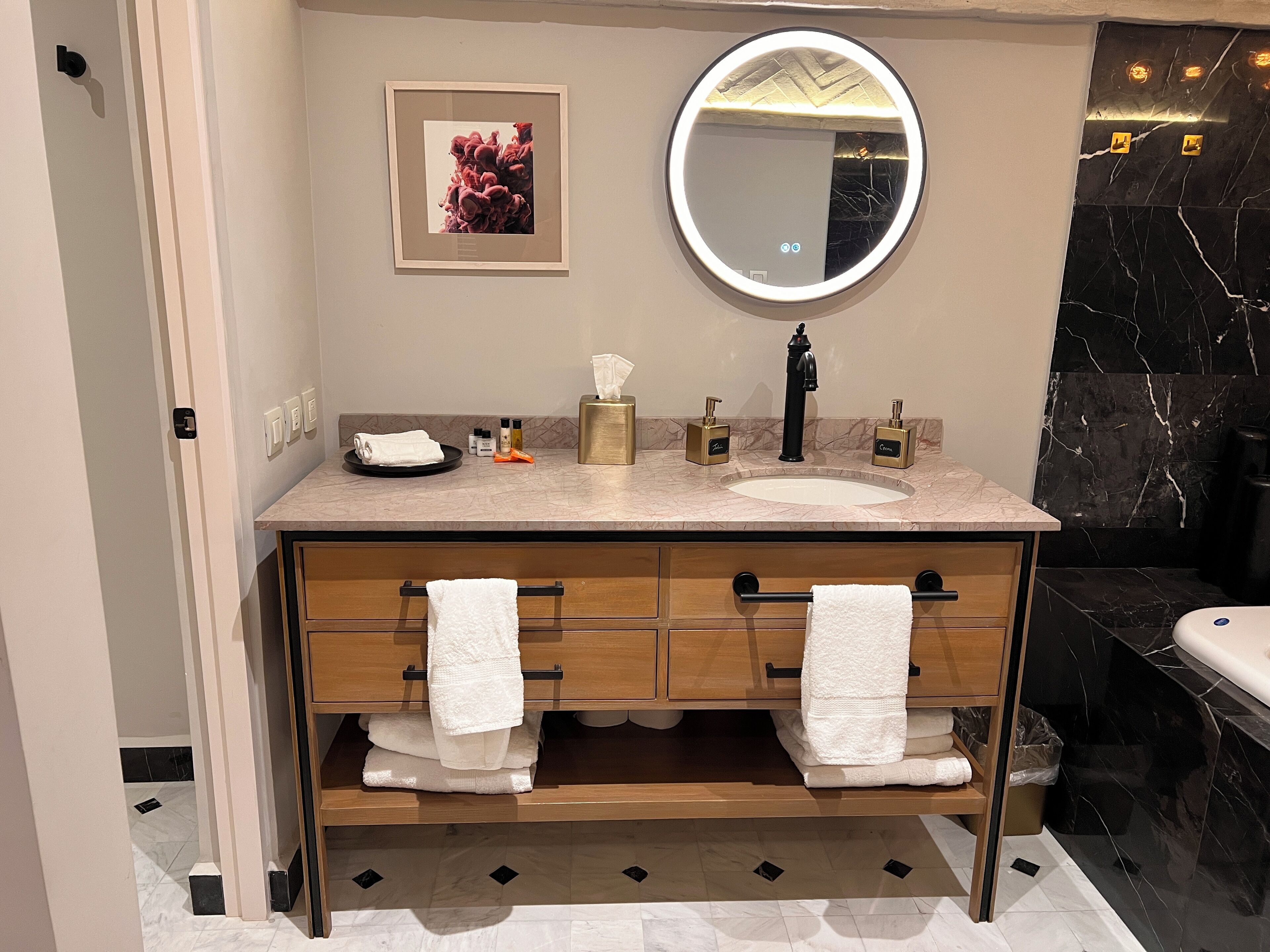 A modern bathroom with a marble sink, a round mirror, a framed artwork, and a towel rack.