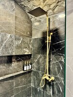 Un baño con cabina de ducha de mármol negro, detalles dorados y un estante con artículos de aseo.
