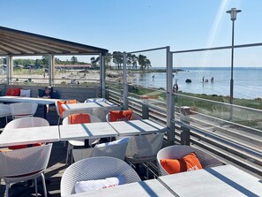 An outdoor seating area with tables and chairs, overlooking a beach and the sea.
