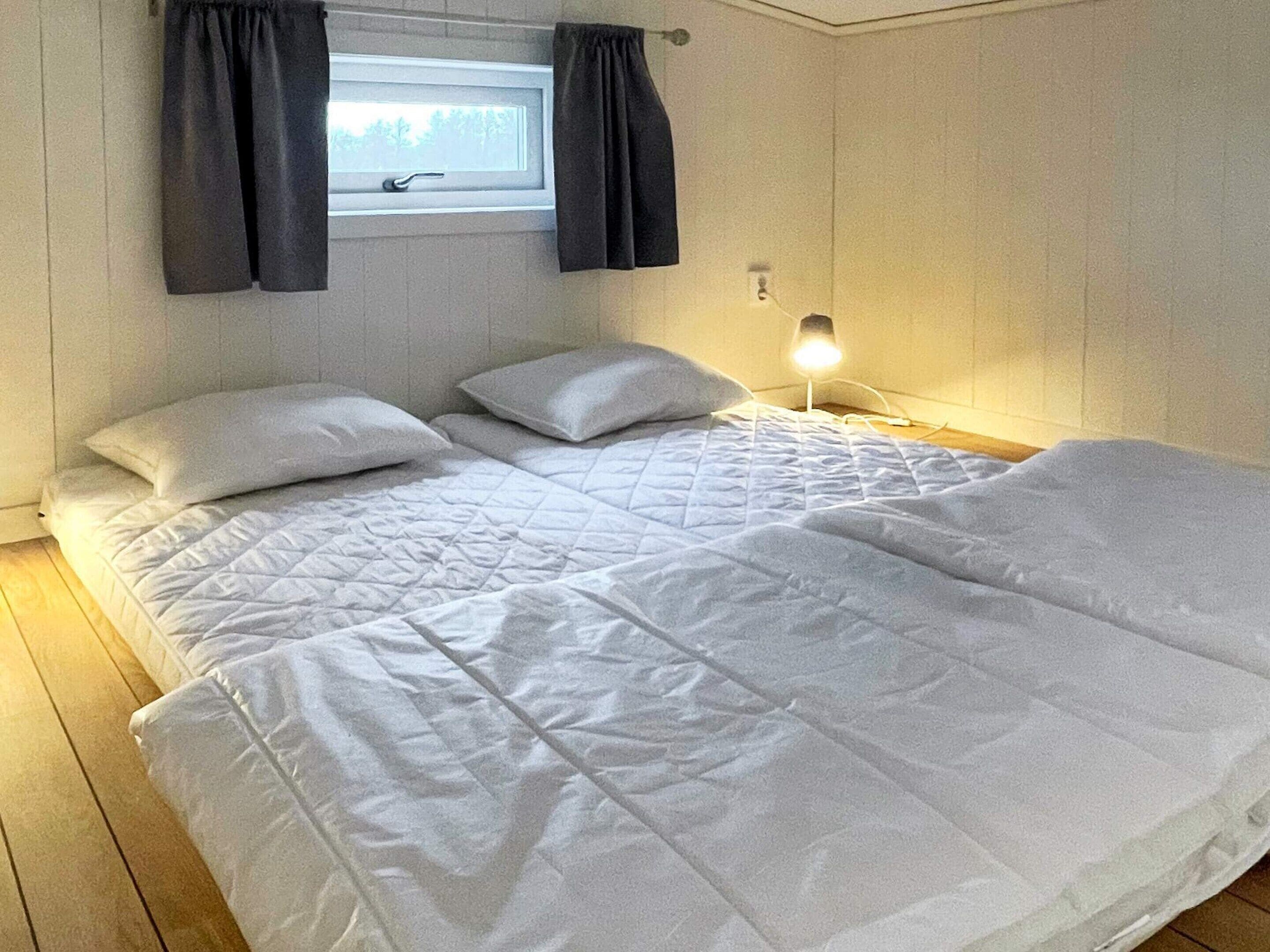 Uma cama de casal com roupa de cama branca, uma mesinha de cabeceira com abajur e uma janela com cortinas.