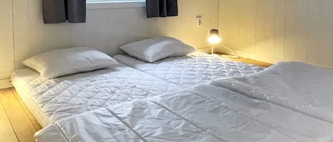 Uma cama de casal com roupa de cama branca, uma mesinha de cabeceira com abajur e uma janela com cortinas.