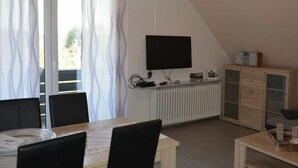 Una habitación con una mesa de comedor, sillas, un televisor montado en la pared, un radiador y un armario con cajones.