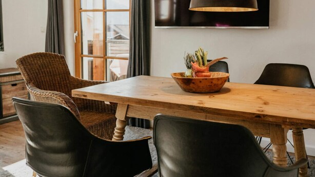 A dining area with a wooden table, black chairs, a woven chair, a bowl with vegetables, a pendant light, and a TV on the wall. - Apartment in Bad Hindelang-bad Oberdorf (Bad Hindelang)