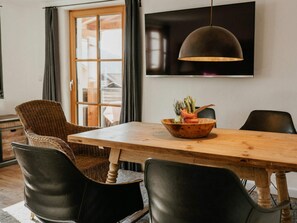 Un coin repas avec une table en bois, des chaises noires, une chaise en osier, un saladier rempli de légumes, une suspension et un téléviseur fixé au mur.