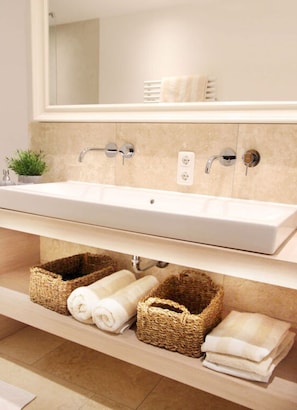 Une salle de bain avec un long lavabo blanc, deux porte-serviettes et des paniers en osier placés sous le lavabo.
