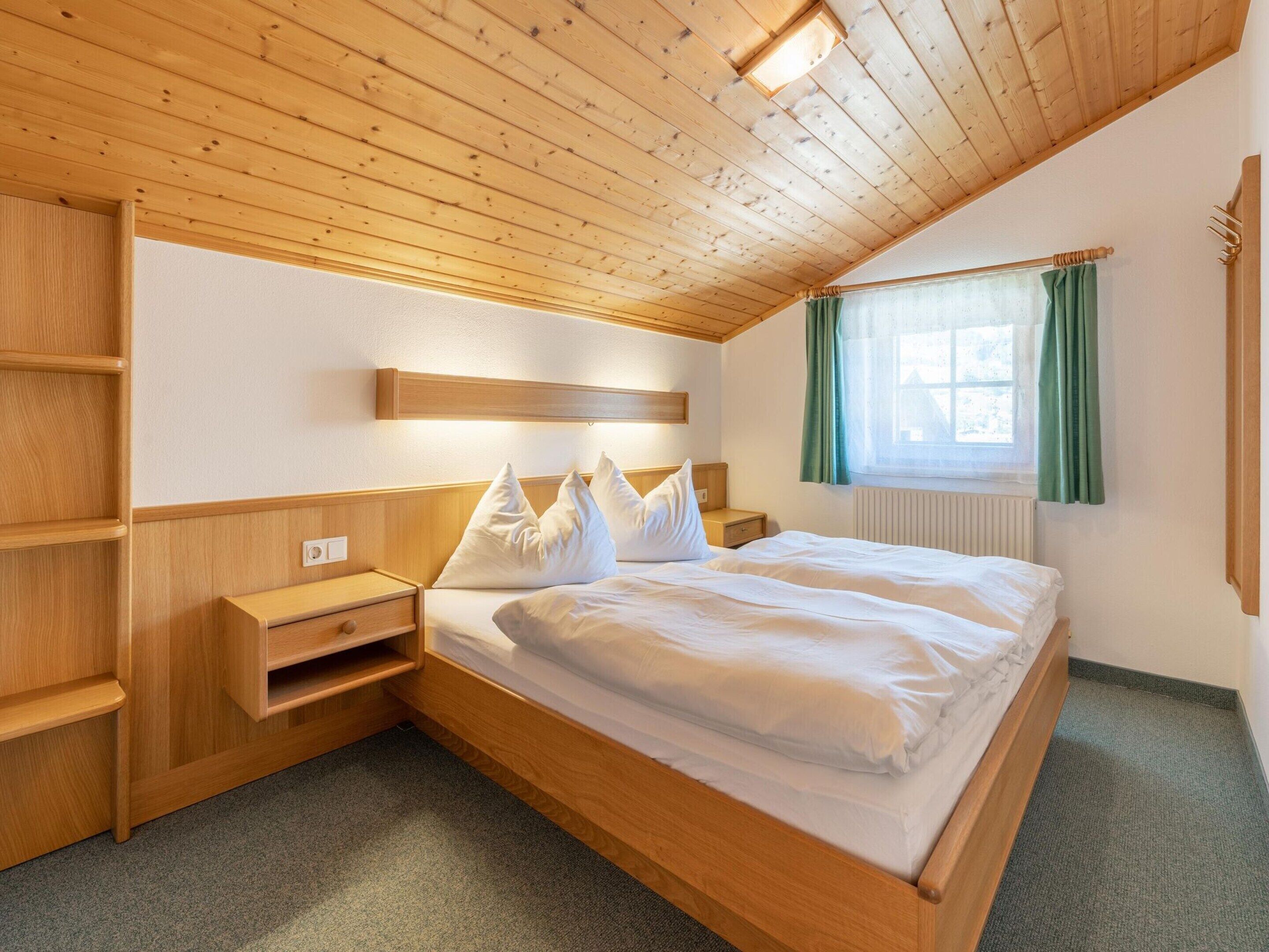 A bedroom with a wooden ceiling, a bed with white bedding, a nightstand, a shelf unit, and a window with green curtains.