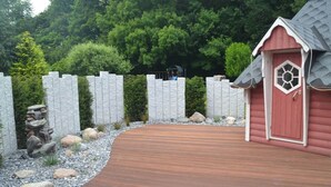A wooden deck leading to a red building with a decorative door, surrounded by a stone wall and a stone waterfall feature. - Saupe Comfortable Holiday Residence (Schaprode)