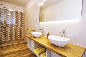 Ein modernes Badezimmer mit zwei großen weißen Waschbecken, einem Holz-Waschtisch und einer gefliesten Duschwand.