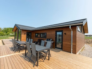 A modern wooden cabin with a deck, a dining area with chairs and a table, and a grill.