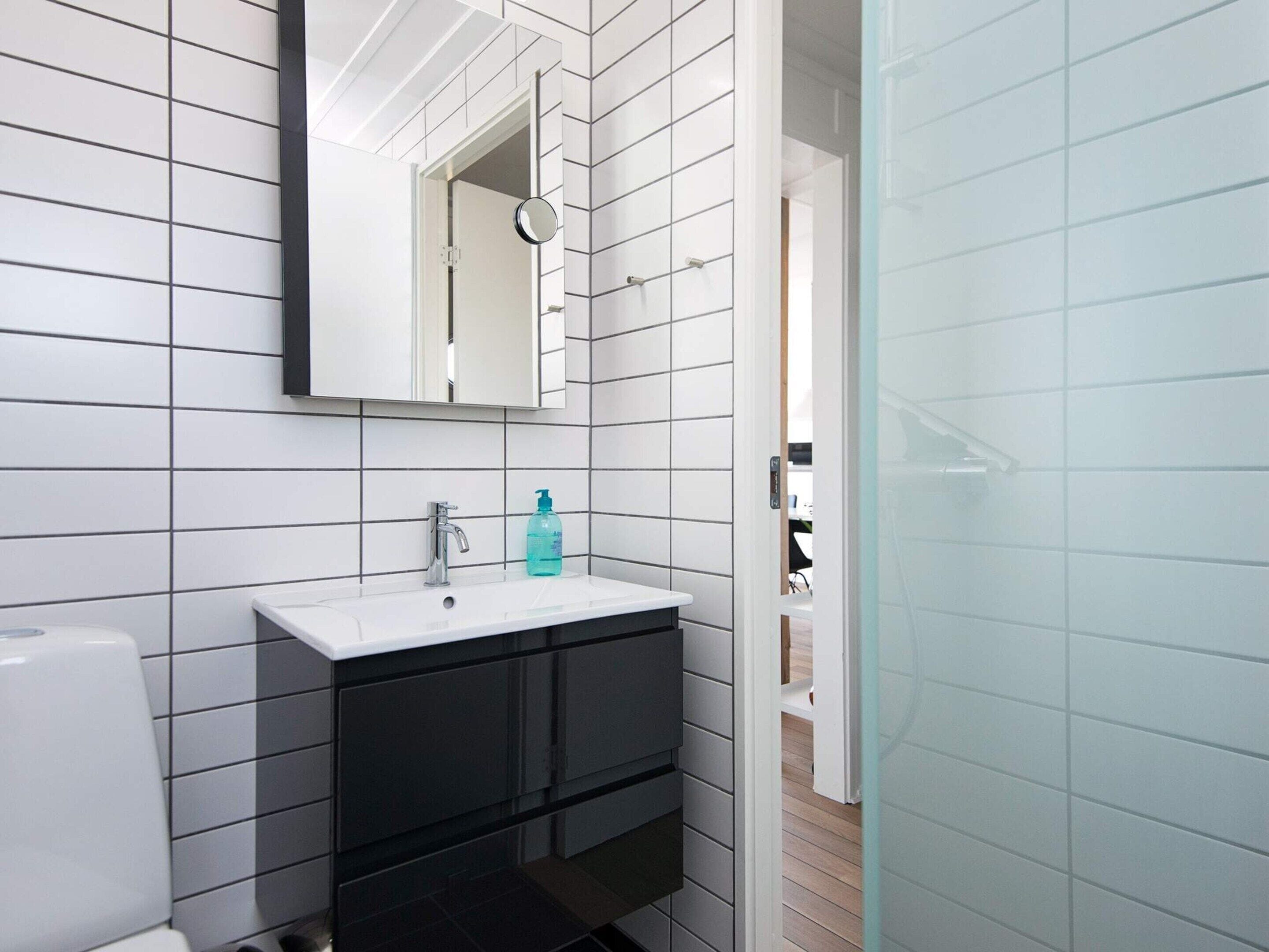 Een badkamer met witte betegelde wanden, een wit wastafelmeubel en zwarte kastjes, met een spiegel.