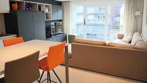 A modern living room with a sofa, a dining table, and chairs. There is a kitchen area with cabinets and a window overlooking a balcony. - Apartment in Nieuwpoort Near Sandy Shores (Nieuwpoort)