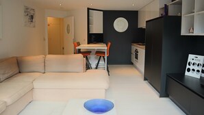 A modern living room with a beige sofa, a dining table with two chairs, and a kitchen area with dark cabinetry. - Apartment in Nieuwpoort Near Sandy Shores (Nieuwpoort)