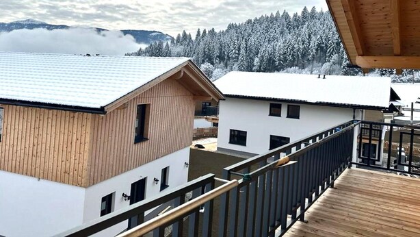 A wooden deck with a view of two buildings, one with a sloped roof and another with a flat roof, set against a snowy landscape with mountains in the background. - Chalet Near Lake Pressegger and Nassfeld ski Area (Hermagor-Pressegger See)