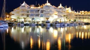 A marina with multiple yachts docked, a large white building with multiple balconies, and a calm water surface reflecting the building and yachts. - Apartment With Beach View (Benalmádena)