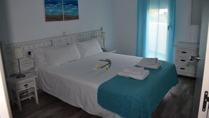 A neatly made bed with white linens, a teal throw blanket, and a pair of folded towels. There is a nightstand with a lamp, a painting of the ocean on the wall, and a window with blinds. - Apartment With Beach View (Benalmádena)