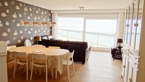 A dining area with a table and chairs, a sofa, and a view of the sea through large windows. - Attic Apartment Nieuwpoort by the Sea Wall (Nieuwpoort)