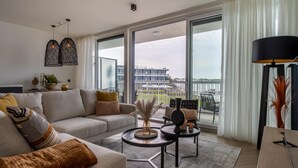 Ein modernes Wohnzimmer mit Sofa, Couchtisch und Sesseln, große Fenster mit Blick auf ein Gebäude und ein Gewässer.