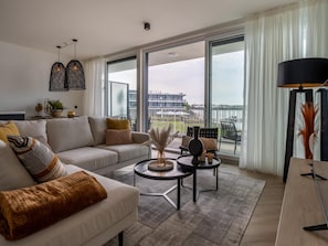 A modern living room with a sofa, coffee table, and chairs, large windows with a view of a building and a body of water. - Apartment in Zeeland With Marina and Views (Arnemuiden)