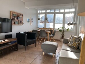 Ein modernes Wohnzimmer mit Flachbildfernseher, bequemen Sitzgelegenheiten, Essbereich und großen Fenstern mit Blick ins Freie.