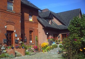 A red brick building with a steeply pitched roof, a front porch with steps, and a garden with various flowers and plants. - Holiday Apartment Kirchhoff (Luetjensee)