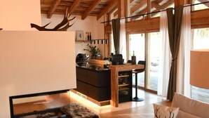 Un salon moderne avec une cheminée, un canapé agrémenté d’un coussin à motif de cerf, et un coin cuisine avec des poutres en bois et de grandes fenêtres.