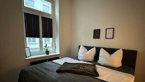 Une chambre avec un grand lit, une fenêtre avec des stores, une plante en pot et deux tableaux encadrés accrochés au mur.