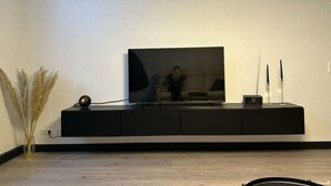 Un salon moderne avec un téléviseur à écran plat fixé sur un meuble noir, un vase contenant des plantes séchées et deux bougies posées sur le meuble.