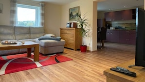 A modern living room with a sofa, a coffee table, and a red rug. There is a wooden cabinet with a plant, and a kitchen area with cabinets and a refrigerator. - Ottenhain Natural Paradise (Kottmar)