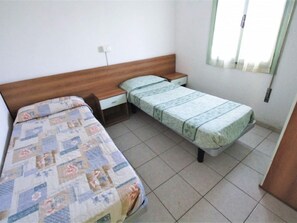 A small, simple bedroom with two single beds, a wooden headboard, a bedside table, and a window with blinds.