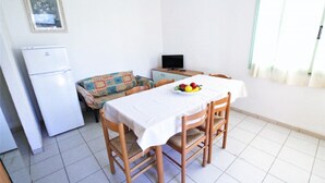 Un comedor con una mesa dispuesta para cuatro personas, un televisor sobre un mueble bajo, un refrigerador y un sofá estampado.