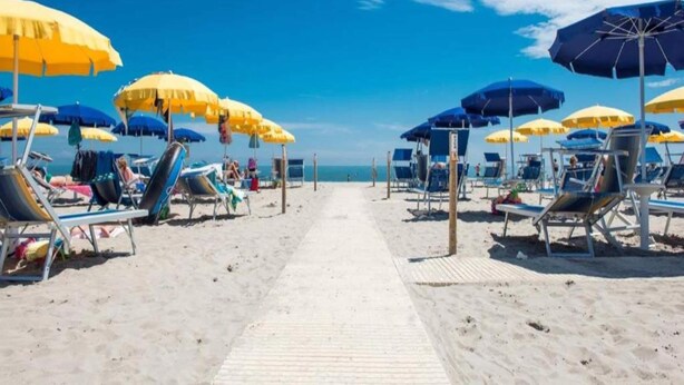 Una playa con filas de tumbonas y sombrillas, un paseo marítimo de madera y el océano al fondo.