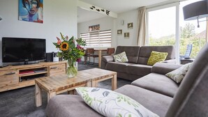 En moderne stue med et fladskærms-tv, et træ sofabord, en grå sofa og en vase med blomster.