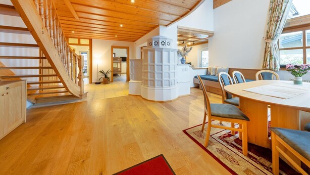 Un amplio salón con escalera de madera, zona de estar central con mesa, y vistas al exterior a través de grandes ventanales.