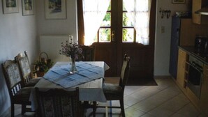 Un coin repas avec une table dressée pour deux personnes, un vase rempli de fleurs et une vue sur l’extérieur à travers une porte ornée de rideaux.