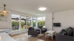 Un salón moderno con un sofá, sillones, una mesa de centro y un televisor montado en la pared. Una ventana grande ofrece vistas al exterior.