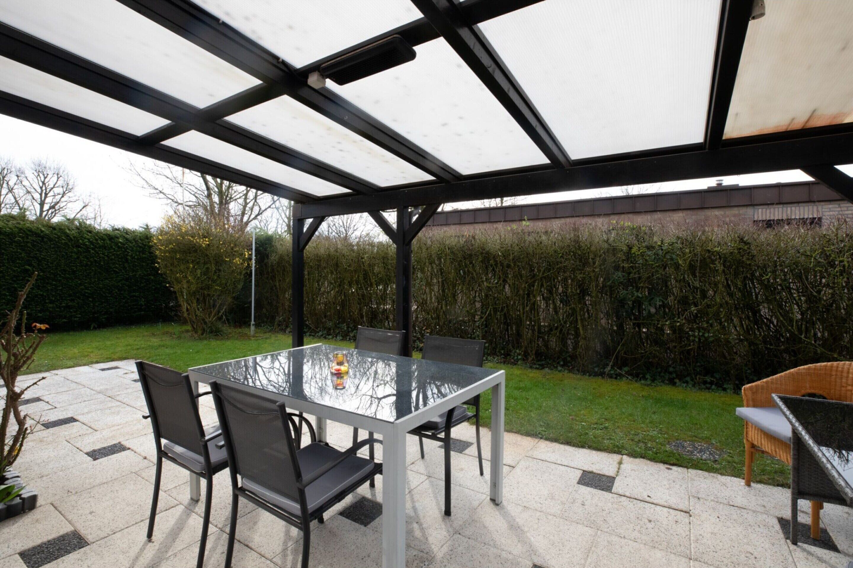 An outdoor patio area with a glass table and chairs, surrounded by a metal frame and covered with a translucent roof.