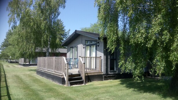 A small cabin with a deck, surrounded by trees and a well-maintained lawn. - Cottage in Brookland Near Romney Marsh Beach (Romney Marsh)
