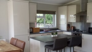 A modern kitchen with white cabinets, a central island with bar stools, a window with a view, and a stainless steel range hood. - Cottage in Brookland Near Romney Marsh Beach (Romney Marsh)