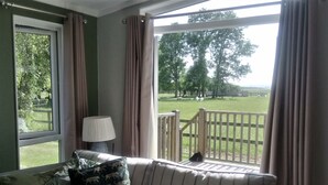 A room with a sofa, a lamp, and a view of a grassy area with trees and a wooden deck. - Cottage in Brookland Near Romney Marsh Beach (Romney Marsh)